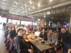 Indoor cafe/bar with long table, seating 17 delegates from IHEC conference, all face the camera smiling