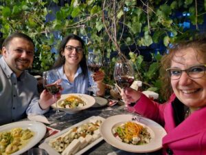 AECED colleagues Susanne and Kardelen with IHEC colleague, seated at outdoor round table with food and wine, smiling.