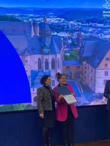 Prof Susanne Weber from Marburg university with certificate at IHEC conference, next to IHEC colleague. The slide behind on the large screen is a scenic view of Marburg.