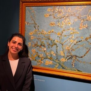 Kardelen Dilara Cazgir stands in front of Vincent Van Gogh's almond blossom painting, which is in a gold frame. She smiles. Her dark hair is tied back and she wears a while top with a dark suit jacket over.