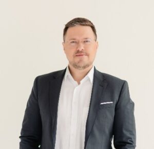 Uldis Dumins stands in front of a white background, dressed in a white shirt and grey suit. He has short light brown hair and a slight beard and moustache. He wears glasses and has an approachable expression on his face.